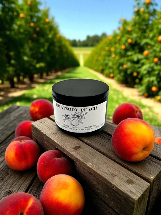 A black candle tin labeled Rhapsody Peach resting on a crate surrounded by peaches with peach trees in the background.
