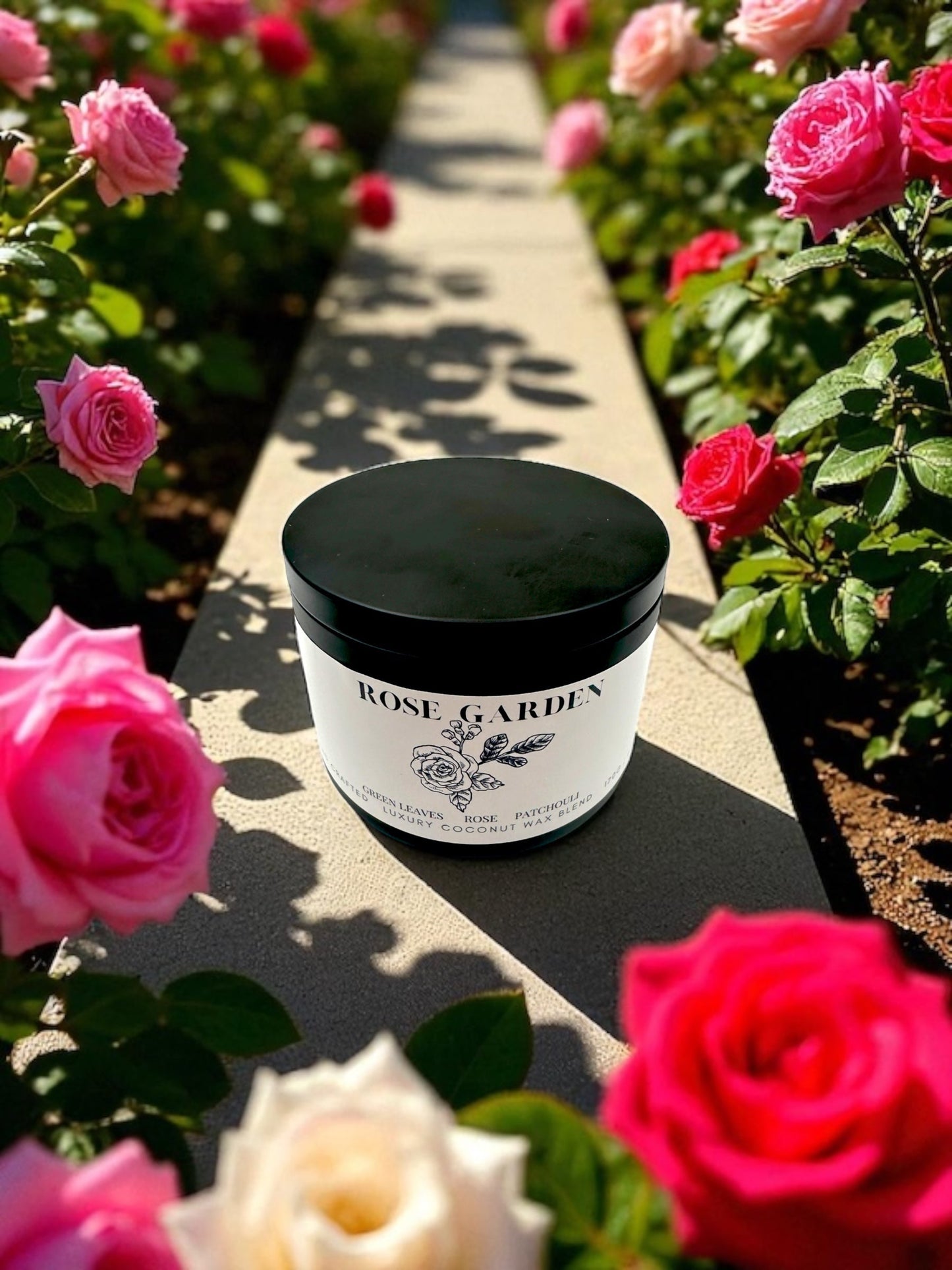 A black candle tin labled Rose Garden  resting on concrete ledge surrounded by rose bushes. 