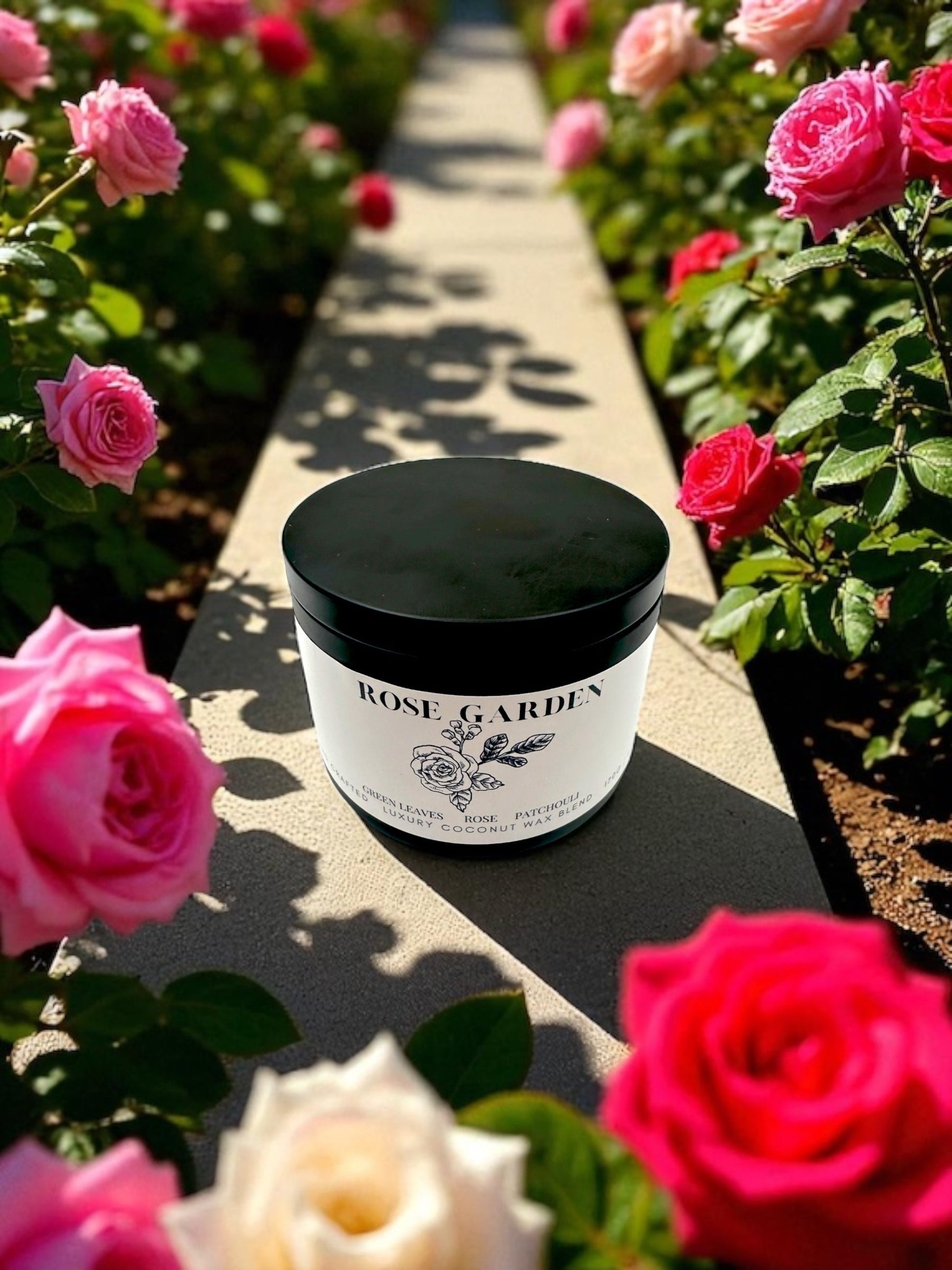 A black candle tin labled Rose Garden  resting on concrete ledge surrounded by rose bushes. 
