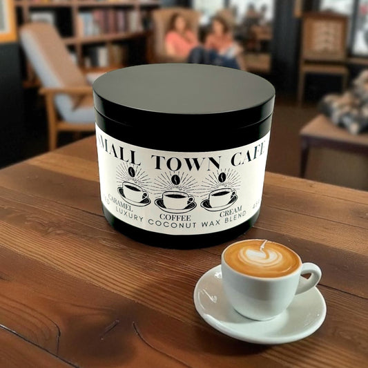 A black candle tin labeled Small Town Cafe on a wooden table with a hot late with the background of a small cozy coffee shop.
