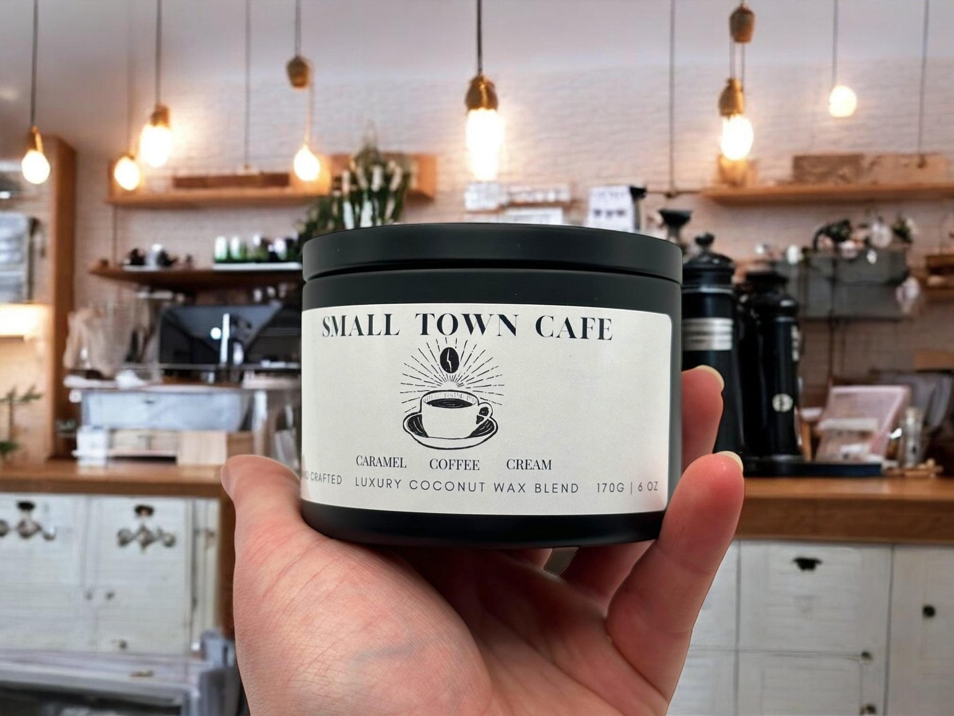 A hand holding a black candle tin labeled small town cafe with a coffee shop in the background.