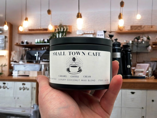 A hand holding a black candle tin labeled small town cafe with a coffee shop in the background.