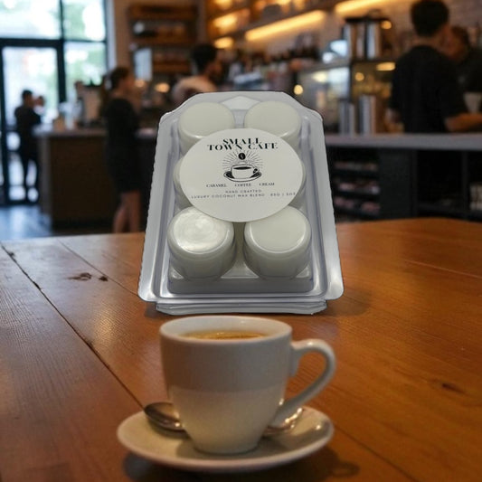 A clear wax melt clamshell labeled Small Town Cafe on a wooden table with a cup of coffee and a coffee shop in the background.