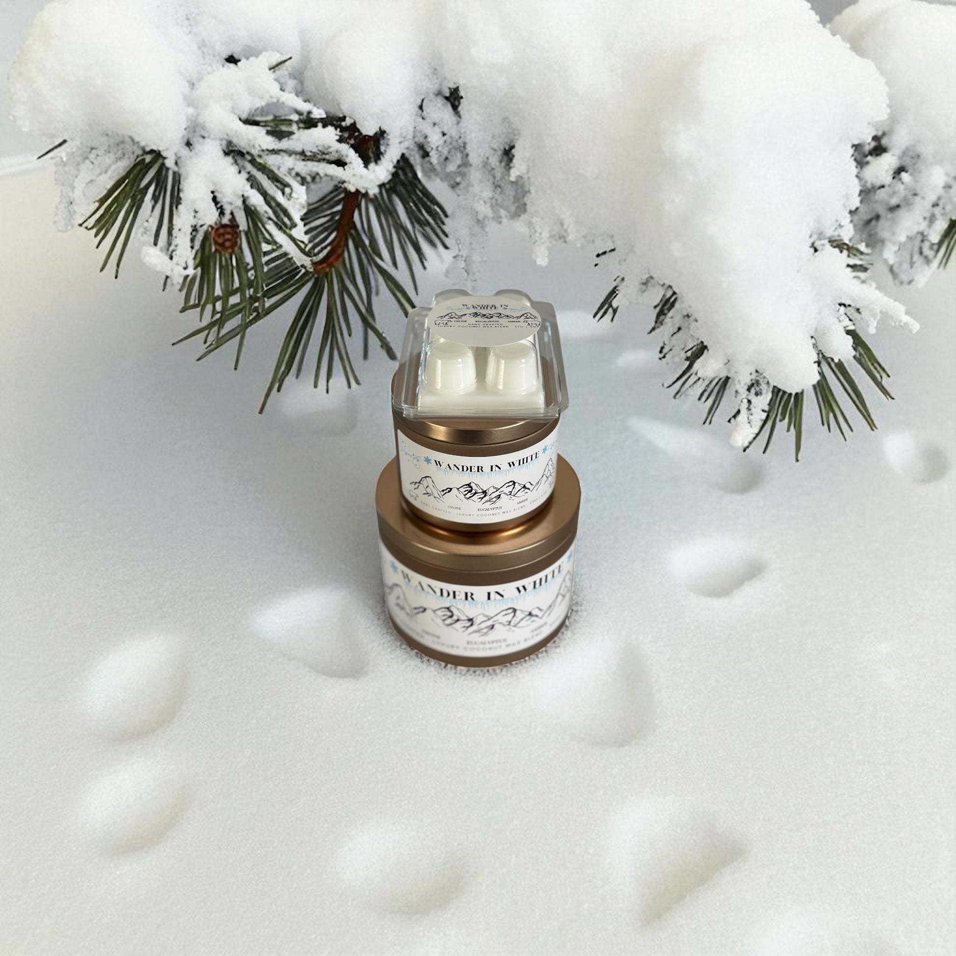 A collection of Gold candle tins and white wax melt labeled "Wander in White" on fresh snow and a snow covered pine tree branch in the background.
